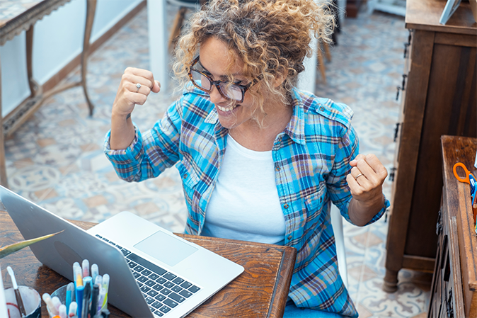 Young woman pumping her arms in excitement as she sits in front of a laptop